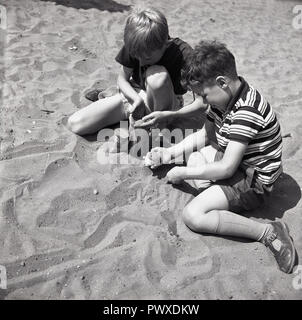 1950, historische, zwei junge Männer in T-Shirts, Shorts und Sandalen sitzen zusammen an einem Sandstrand, England, UK. Stockfoto