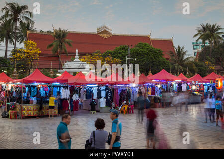 Menschen und viele Stände voller Kleider an der berühmten Nachtmarkt in Vientiane, Laos, in der Dämmerung. Wat Chanthaburi (Chanthaboury Tempel im Hintergrund ist. Stockfoto