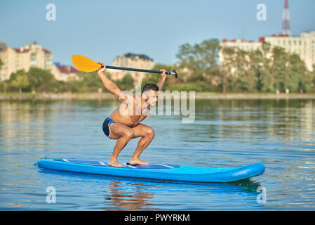 Portrait von Sportsman Holding lange Ruder über Kopf, Schwimmen auf Paddle Board in Stadt See. Schöner Junge mit fit Körper und starke Muskeln, Training während der sonnigen Sommertag. Konzept der gesunden Lebensweise. Stockfoto