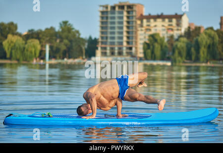 Hübscher junger Boy tun Hand stehend auf paddle Board während der heißen Sommertag. Sportler mit perfekter Figur, sportlichen Körper, trägt blaue Shorts, Ausbildung außerhalb. Konzept der gesunden Lebensweise. Stockfoto