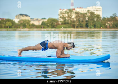 Schöne sportliche Mann tun plank Übung auf paddle Board an Stadt See. Heiße Junge mit perfekter Passform Abbildung, sportliche Figur, trug in blauen Shorts, Ausbildung Yoga outdoor. Konzept der gesunden Lebensweise. Stockfoto