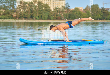 Muskulöse mann Üben Yoga, Balance Körpergewicht, Schwimmen auf Paddle Board in der Mitte der Stadt, See, während der sonnigen Sommertag. Sportler mit perfekter Figur, trägt blaue Shorts, Ausbildung außerhalb. Stockfoto