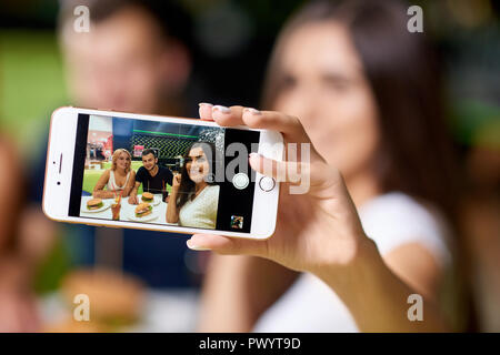 Selektiver Fokus der Mädchen halten Telefon und unter selfie mit Freunden im Cafe. Unternehmen am Tisch sitzen, Telefon, lächeln und posieren beim Essen lecker fast food. Konzept der Freundschaft. Stockfoto