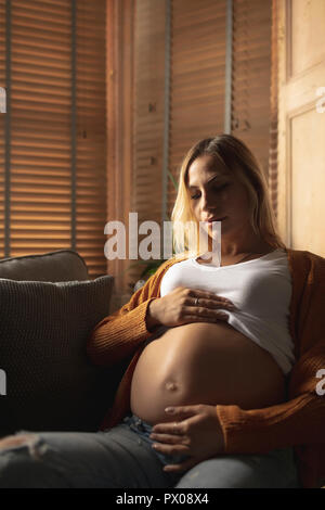 Schwangere Frau sitzt auf einem Sofa ihr Bauch berühren. Stockfoto