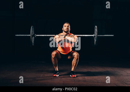 Schöne muskulöse Mann Kniebeugen mit Langhantel in dunklen Fitnessraum Stockfoto