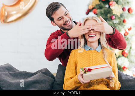 Schöner Mann schließen die Augen und lassen Sie sich überraschen mit Geschenkbox für die Frau zu Hause mit Weihnachtsbaum Stockfoto