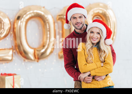 Schönen lächeln Ehepaar in Santa Hüte umarmen zu Hause mit 2019 neuen goldenen Luftballons Stockfoto