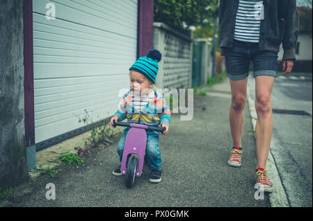 Eine Mutter ist zu Fuß auf der Straße neben ihr Toddler, ist Reiten ein Dreirad Stockfoto