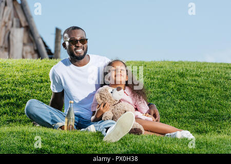 Happy african american Vater und Tochter sitzen auf Hügel mit Soda und Teddy Bär am Vergnügungspark Stockfoto