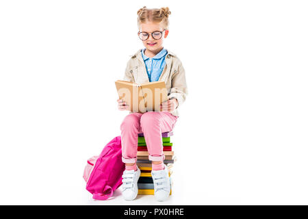 Adorable Schülerin lesen Buch sitzend auf Stapel Bücher mit Rucksack auf weissem, Bildung Konzept isoliert Stockfoto