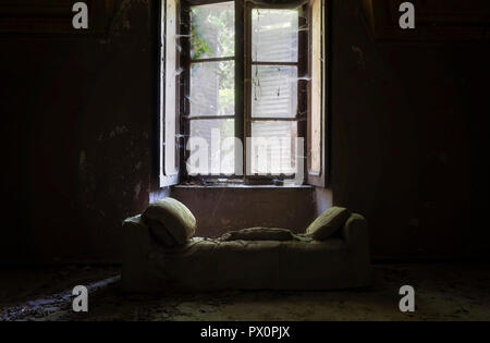 Innenansicht mit einem Sofa vor dem Fenster in einem dunklen Raum eines verlassenen Hauses in Italien. Stockfoto