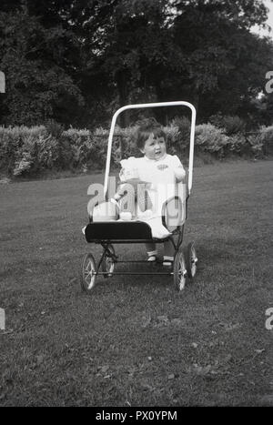 1950, historische, kleine Mädchen im Freien in einem Park mit ihren weichen Spielzeug in Kinderwagen ihres Metall Puppe oder Spielzeug Kinderwagen, England, UK. Stockfoto