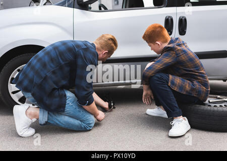 Vater mit dem Wagenheber anheben Auto für Reifenwechsel, Sohn sitzen auf Reifen Stockfoto