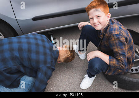Vater mit dem Wagenheber anheben Auto für Reifenwechsel, glücklich Sohn an der Kamera auf der Suche Stockfoto