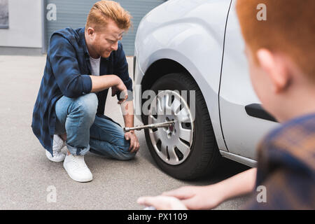 Vater Reifenwechsel im Auto mit Radschlüssel, Sohn sitzen. Stockfoto