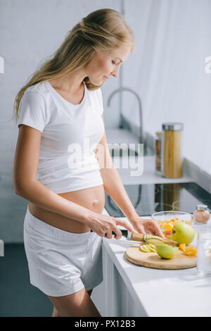 Seitenansicht der schwangeren Frau mit Messer kochen Obstsalat zu Hause Stockfoto