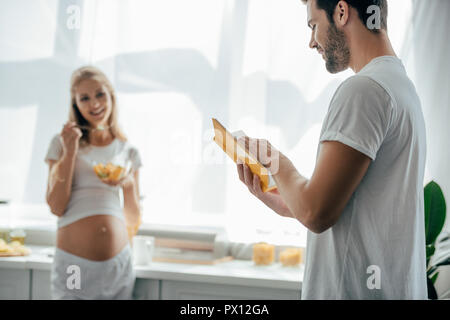 Selektiver Fokus der schwangeren Frau mit Obst und Salat Mann mit Buch in der Küche zu Hause. Stockfoto