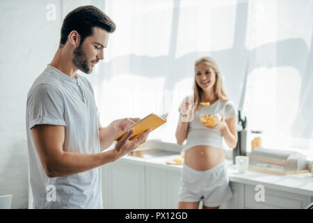 Lächelnd schwangere Frau mit Obst und Salat Mann mit Buch in der Küche zu Hause. Stockfoto