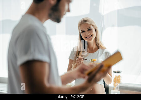 Selektiver Fokus der lächelnden schwangere Frau mit Obst und Salat Mann mit Buch in der Küche zu Hause. Stockfoto