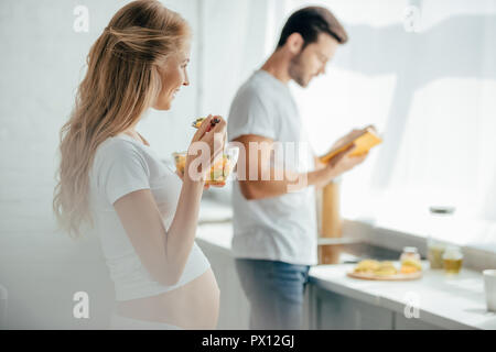 Selektiver Fokus der schwangeren Frau Obst essen Salat, während Mann mit Buch stehen an der Theke in der Küche Stockfoto