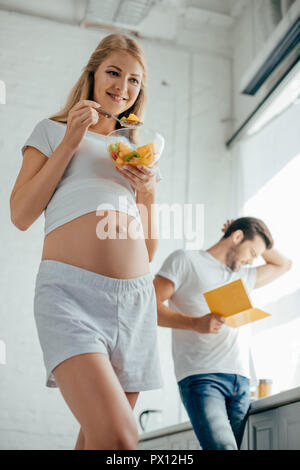 Selektiver Fokus der lächelnden schwangere Frau Obst essen Salat, während Mann mit Buch stehen an der Theke in der Küche Stockfoto