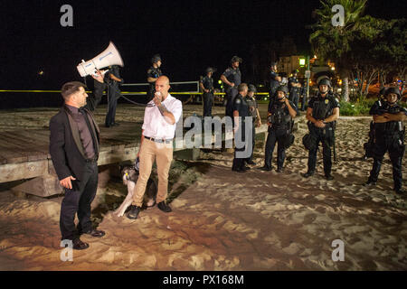 Wie die Polizei in harte Hüte hören, eine Linke-Demonstrator von Hus Hund begleitet spricht auf eine politische Kundgebung am Hauptstrand in Laguna Beach, CA. Stockfoto