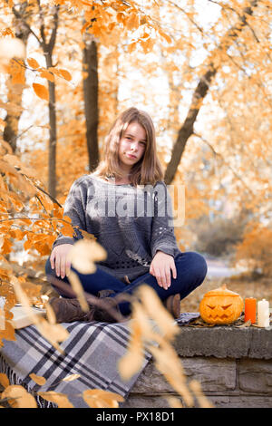 Schöne jugendlicher Mädchen im Herbst Garten saß auf dem Zaun, auf der Wolldecke plaid Decke mit Halloween Kürbis und Kerzen Stockfoto