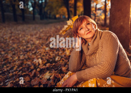 Frau mittleren Alters holding Blumenstrauß aus Ahorn Blätter. Dame genießen Herbst Natur und entspannter sitzen auf dem Boden Stockfoto