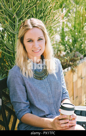 Junge blonde Mädchen sitzt auf einer Bank mit Kaffee Stockfoto