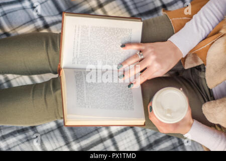 Frau mit Buch auf Beinen und Kaffee in der Hand. Mädchen lesen Buch über retro Decke. Herbst Konzept Bild. Close Up, selektiver Fokus Stockfoto