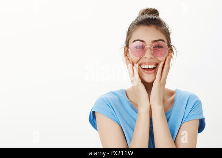 Studio shot von Angeregten gerne schöne Frauen in stilvollen Rosa runde Gläser, berühren Wangen mit Palmen und breit grinsend, erfreut und überrascht, erstaunt und fasziniert über graue Wand Stockfoto