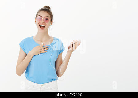 Mädchen zu Fuß auf der Straße treffen Freund. Portrait von positiv überrascht, attraktive Frau in rosa Sonnenbrille und t-shirt, holding Smartphone- und Ohrhörer, berühren die Brust mit Arm, breit grinsend Stockfoto