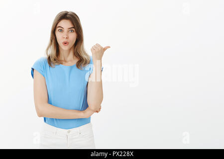 Wow, nicht schlecht. Positive intrigiert attraktive Frau im blauen T-Shirt, Falten und Lippen nach rechts mit Daumen, beeindruckt und neugierig mit der talentierte Musiker an der Partei, diskutieren über graue Wand Stockfoto