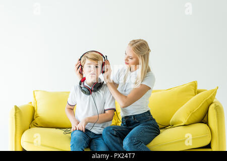 Mutter Kopfhörer tragen am Sohn auf gelben Sofa isoliert auf weißem Stockfoto