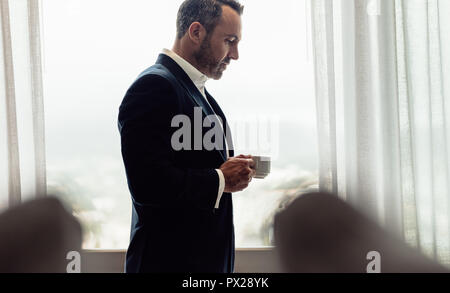Unternehmer stehen im Hotel Zimmer mit einer Tasse Kaffee. Reifer Mann im Anzug trinken Kaffee im Hotel Zimmer am Morgen. Stockfoto