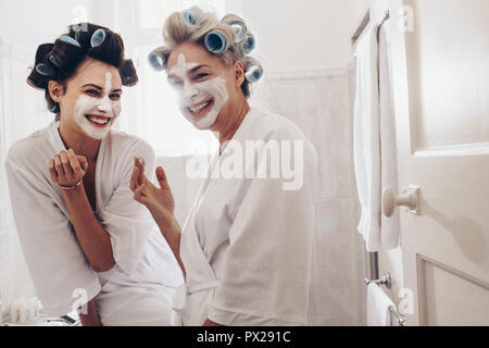 Frauen mit Curling Walzen zu Haar Anwendung Gesichtscreme stehend im Bad. Glückliche Mutter und Tochter Spaß Pflege ihr Gesicht und Haare. Stockfoto