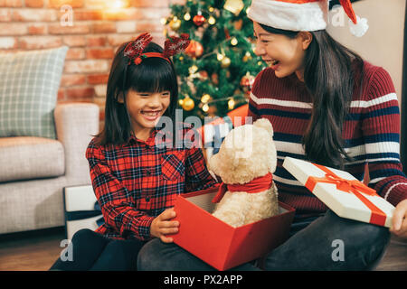 Glückliche Familie, Mutter und Tochter am Weihnachtsbaum zu Hause. Kleines Mädchen mit Reh auf Kopf öffnen Geschenk im Wohnzimmer. junge Mamma senden niedlichen Teddy. Stockfoto