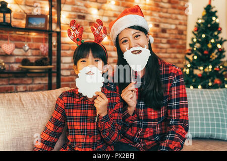 Attraktive santa Mädchen und schöne Kind mit Rotwild auf Kopf Spaß im Wohnzimmer. Junge Mama mit weißen Bart selbst wie Sant zu tun gekleidet Stockfoto