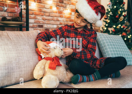 Schöne Weihnachten Kind in Red Hat sitzen auf der Couch und spielen Ihr neues Spielzeug. Mädchen mit ihrem Liebling Teddybär im Wohnzimmer. Xmas Tree und Rot Stockfoto