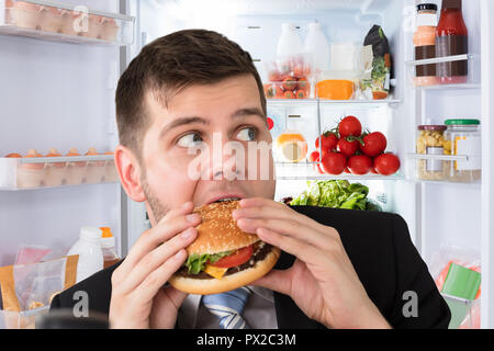 Junge Unternehmer Essen Burger vor dem offenen Kühlschrank Stockfoto