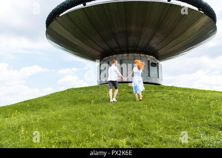 Ansicht der Rückseite des redhead Paar halten sich an den Händen und laufen auf grasbewachsenen Hügel vor modernes Gebäude Stockfoto