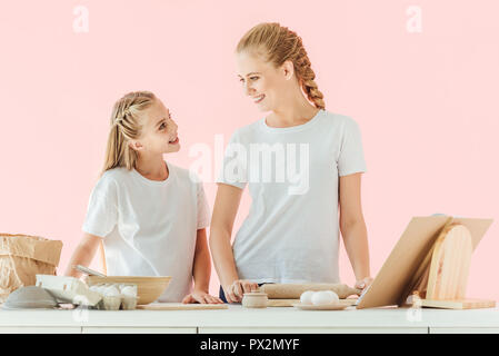 Lächelnde Mutter und Tochter in den weißen T-Shirts sich einander beim Kochen gemeinsam auf rosa isoliert Stockfoto