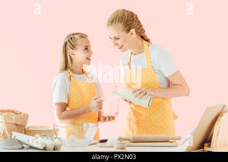 Glückliche Mutter gießen Milch für die Zubereitung von Teig mit Tochter isoliert auf Rosa Stockfoto