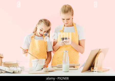 Lächelnde Mutter und Tochter mit Smartphone beim Kochen gemeinsam auf rosa isoliert Stockfoto
