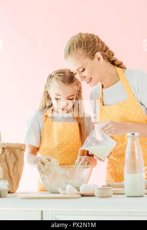 Lächelnde Mutter gießen Milch in den Teig kneten, während Tochter isoliert auf Rosa Stockfoto