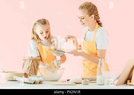 Lächelnde Mutter und aufgeregten kleinen Tochter in Gelb schürzen Zubereitung Teig zusammen isoliert auf Rosa Stockfoto