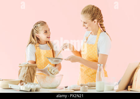 Lächelnde Mutter und Tochter in Gelb schürzen Zubereitung Teig zusammen isoliert auf Rosa Stockfoto