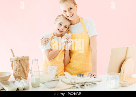 Glückliche junge Mutter und Tochter mit Teig in Form von Herzen, die an der Kamera während der Vorbereitung cookies isoliert auf Rosa Stockfoto