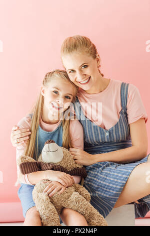 Schöne glückliche Mutter und Tochter mit Teddybär umarmen und mit Blick auf die Kamera isoliert auf Rosa Stockfoto