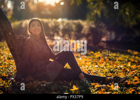Frau im Herbst, Lifestyle Konzept. Eine Asiatische junge erwachsene Frau genießt das sonnige Wetter im Herbst, in einem Park mit Gefallenen gelbe Blätter. Stockfoto